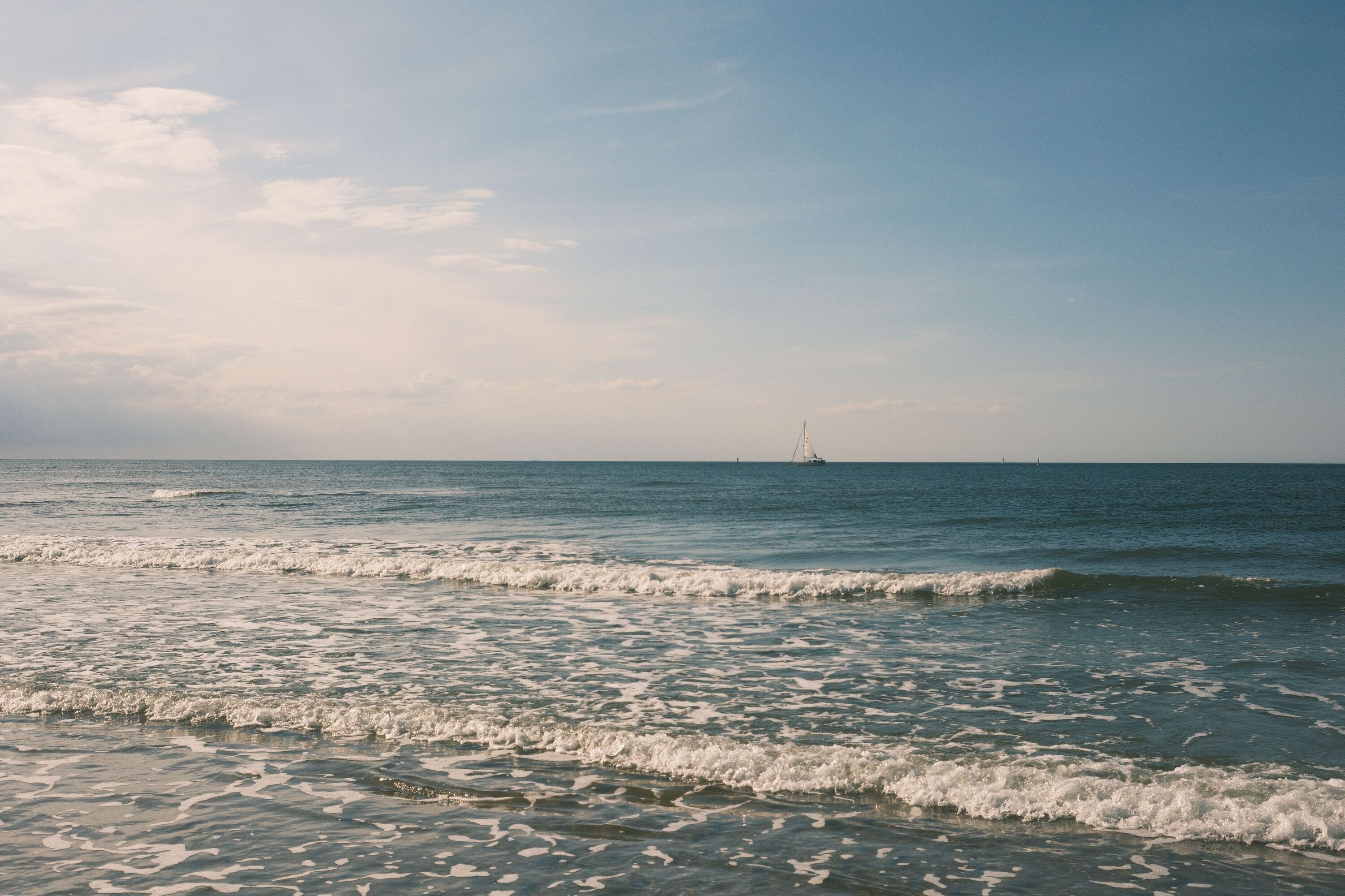 Low waves are lapping towards the shore, a small sailboat is visible on the horizon.