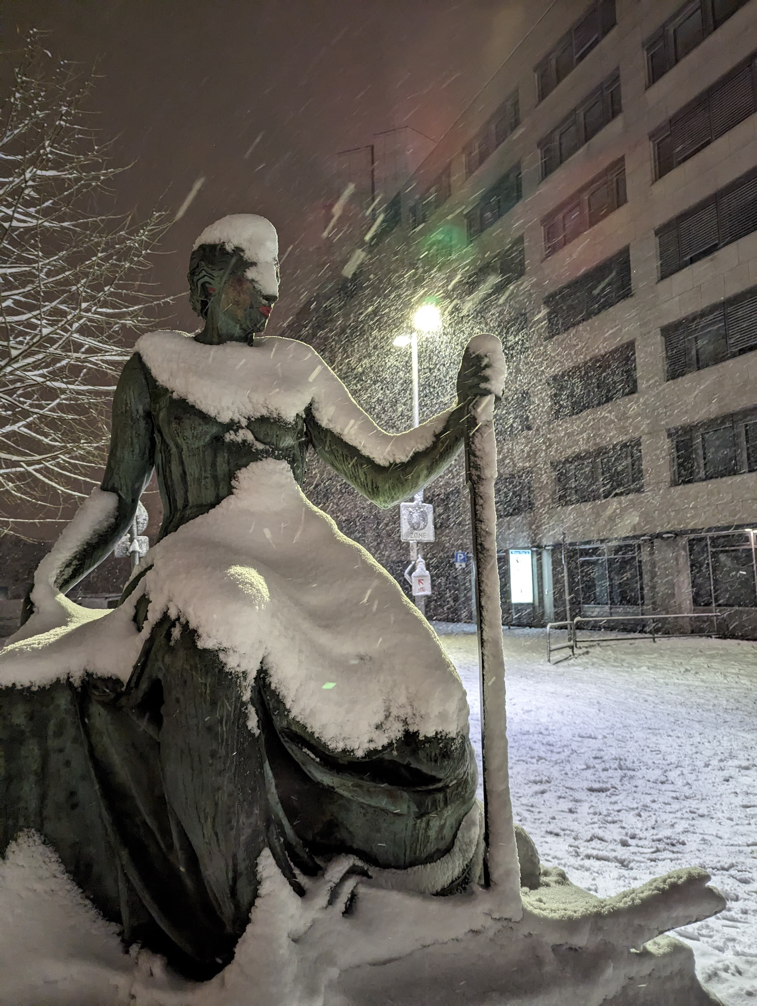 A statue of a woman, partially covered in snow, lit from behind by a street lantern under heavy snowfall.