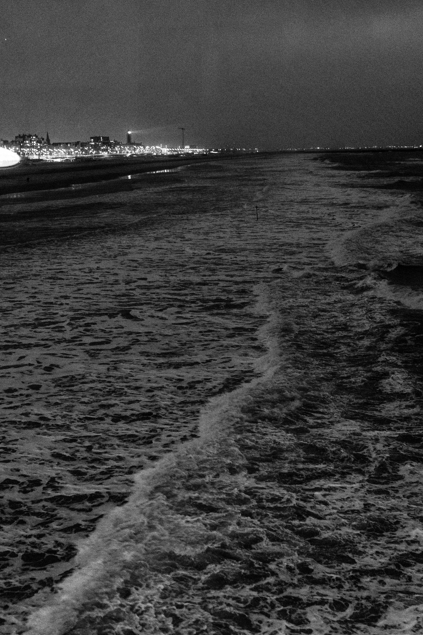 A black-and-white photo of frothy waves approaching the shore, with a lighthouse shining in the background.
